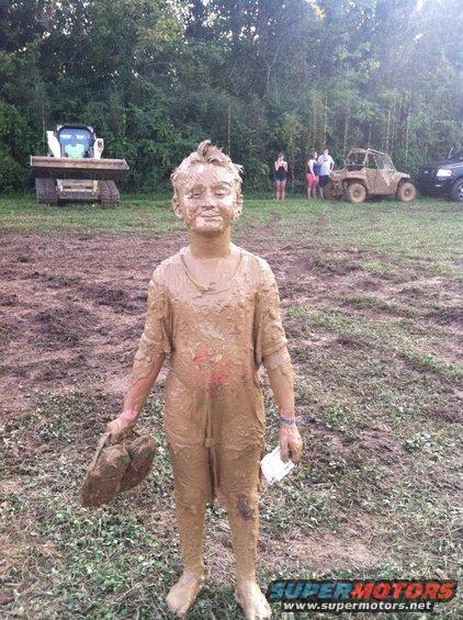 Mud-covered child holding a shoe and paper, standing in a muddy field with a skid steer and ATV in the background