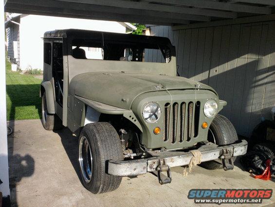 Olive green Jeep CJ-5 with a black soft top, seen from the front at a slight angle under a carport