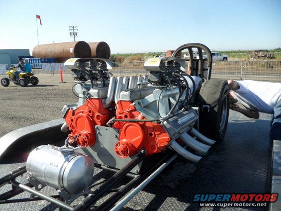 Dragster with dual V8 engines, exposed headers, and large rear tires. A person is working on the rear wheel