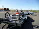 1973 Ford Ranchero - A dragster chassis is loaded onto a trailer, with a gray coupe parked nearby under a clear sky.
