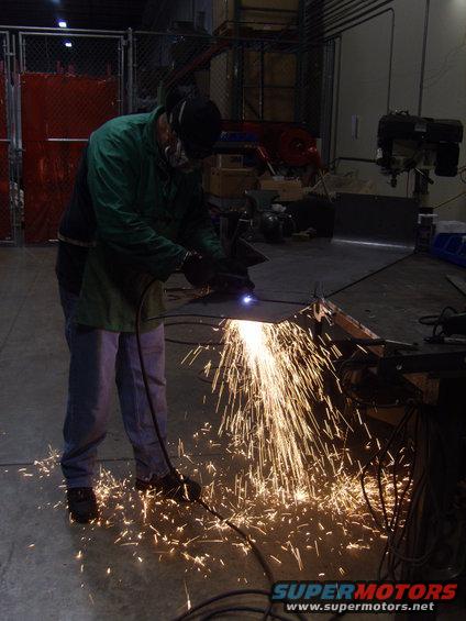 Person in workshop wearing safety gear, cutting a metal sheet with a plasma cutter, creating many sparks