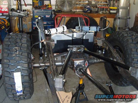 Jeep chassis with large tires, fuel tank, and suspension components in a garage