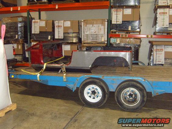 Red and silver Jeep CJ body sections loaded onto a blue trailer, with shelves of boxes in the background