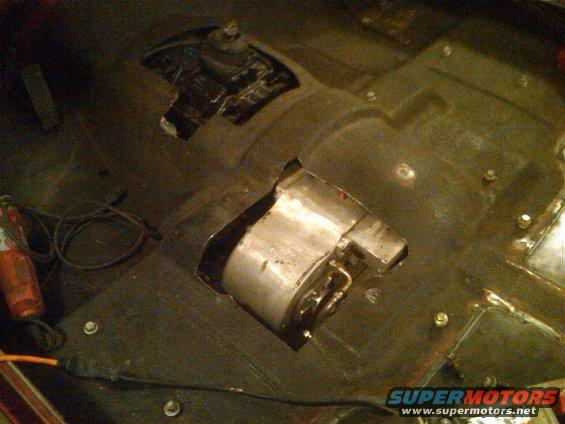 Vehicle floor pan showing cutouts for transmission tunnel and exhaust, with some metal fabrication visible. A power tool and cables are on the left