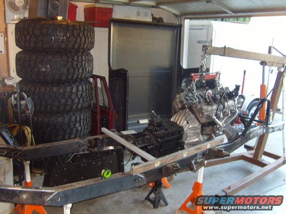 Vehicle frame on jack stands with engine being lowered by a crane. Stack of four large tires to the left