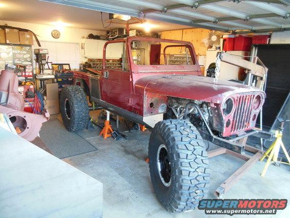 1985 Jeep CJ7 body on jack stands in garage, undergoing restoration. Large off-road tires are mounted