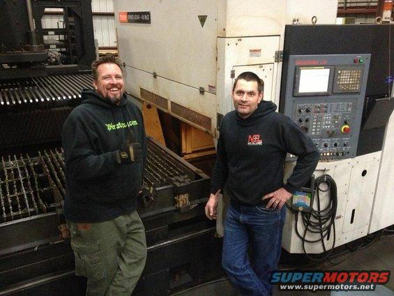 Two men in hoodies stand in front of large industrial equipment with control panels