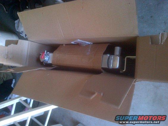 Exhaust pipe with chrome tips and red tag, partially wrapped in brown paper, inside an open cardboard box