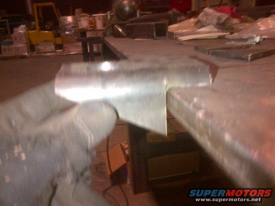 Metal bracket held by a gloved hand, with a workbench and forklift in the background