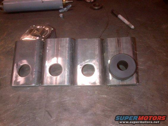 Metal brackets with holes and a rubber bushing sit on a workbench next to a cylinder and a marker