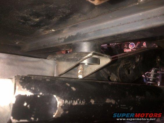 Close-up of a metal bracket with a rubber bushing, part of a vehicle's suspension system. Some text is visible in the background