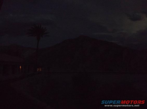 Dark desert landscape with mountains, a palm tree, and a building with lights on