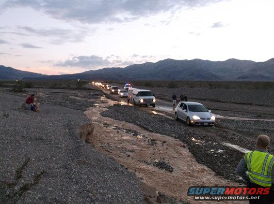Multiple vehicles, including cars and a van, driving in a line through a desert wash with mountains in the background