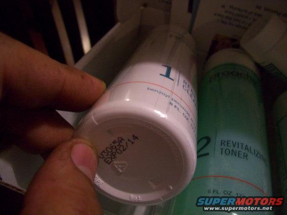 Hand holding a Proactiv Revitalizing Cleanser bottle with expiration date 02/14 visible. Another Proactiv bottle is next to it