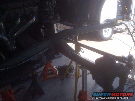 Vehicle frame and suspension parts are supported by jack stands, with a wheel visible in the background