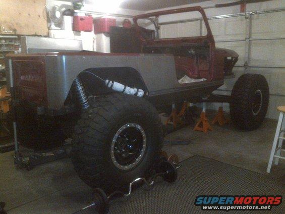 Jeep CJ body on jack stands in garage, missing doors and roof. Large off-road tires are visible