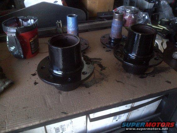 Two black painted automotive hubs with splined bores sit on a cardboard surface next to a red and black can