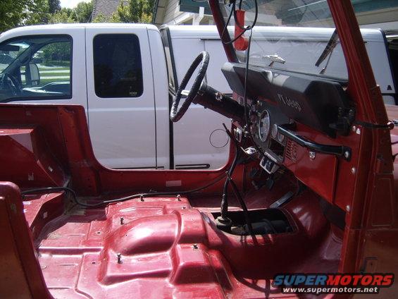 Red Jeep interior with removed seats, showing floor and dashboard. A white truck is parked behind