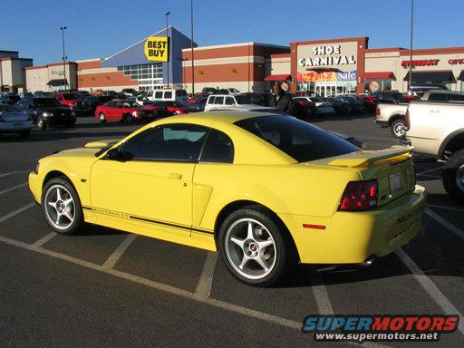 Yellow 2003 Ford Mustang parked in a lot with stores in the background