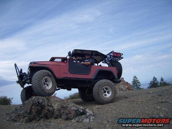 Red Jeep CJ-series off-roading on rocks, driver visible, aftermarket roll cage and spare tire carrier