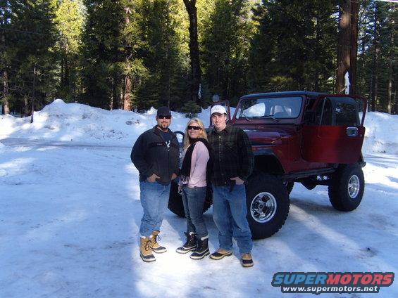 Three people stand in front of a red Jeep CJ in a snowy wooded area