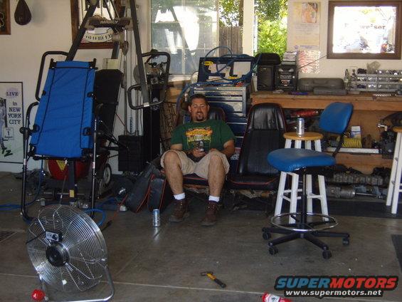 Man sitting in a garage with a fan, chairs, and tools around him