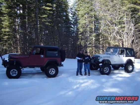 Two people stand between a red Jeep CJ and a white Jeep CJ in a snowy forest. The white Jeep has 'LAREDO' written on its side