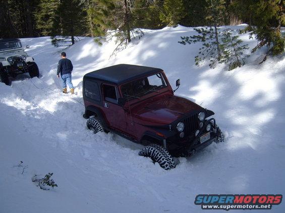Red Jeep CJ-series stuck in snow, black hardtop, winch cable attached to white Jeep in background