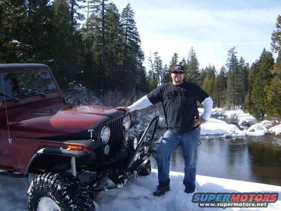 Man in black t-shirt and jeans stands by a red 1985 Jeep CJ7 in a snowy landscape