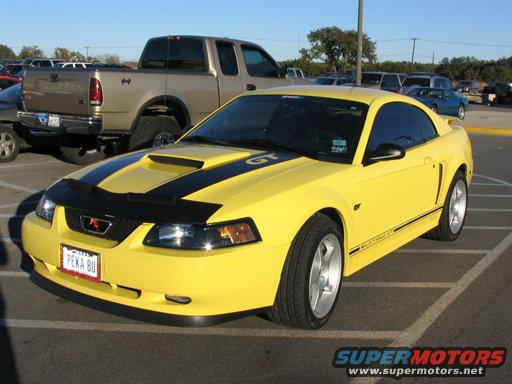 Yellow 2003 Ford Mustang GT with black racing stripes on hood and side. Parked in a lot with a pickup truck behind it
