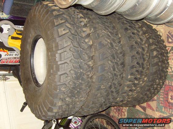 Four off-road tires stacked, one with a white rim, on a carpeted floor