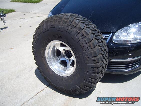 large off-road tire in front of black car's bumper and headlight