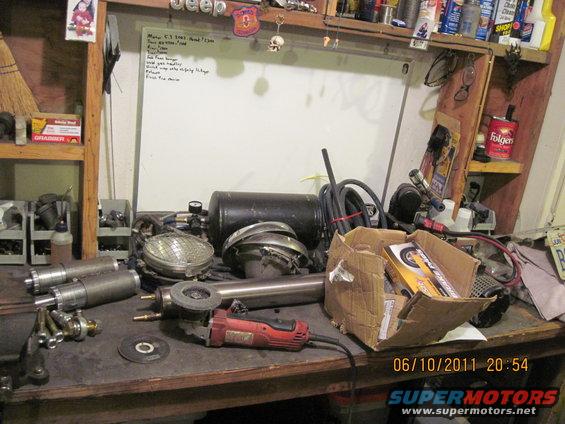 Workbench cluttered with tools including an angle grinder, spare parts like headlights and exhaust pipes, and a box of new components