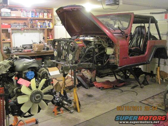 Red Jeep CJ7 with hood open, engine removed, on jack stands in a garage