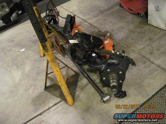 Front axle assembly, possibly from a Jeep, supported by yellow jack stands. A tag is attached to the differential housing