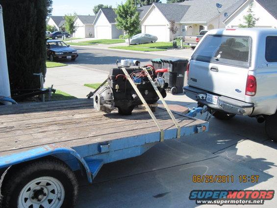 Engine secured with straps on a blue trailer, towed by a silver pickup truck. Houses and trees in background