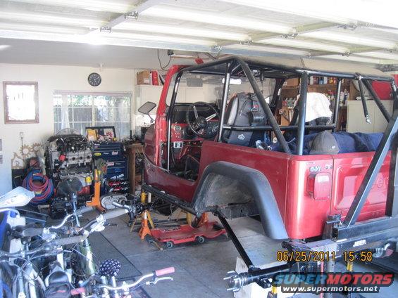 Red Jeep CJ-series body with roll cage, missing doors and fenders, on jack stands in a garage