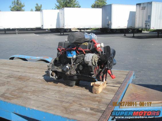 Engine block with fan and alternator on a wooden platform on a trailer. White semi-trailers are in the background