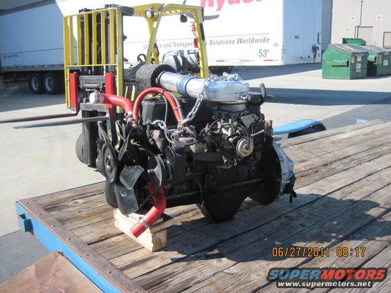 Engine on wooden pallet, supported by wooden blocks. Red hoses, silver exhaust pipe, and chain visible