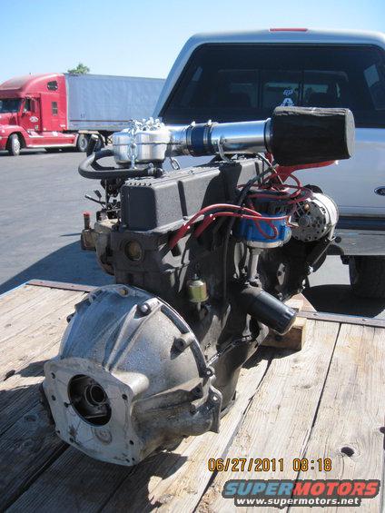 Engine and transmission assembly on a wooden surface, with a semi-truck in the background