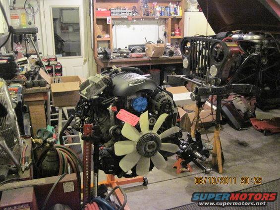 Jeep CJ-series grille and engine on stands in a garage. The grille is black with round headlights. A VORTEC engine is in the foreground