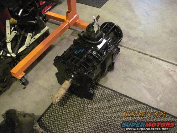 Black transmission with a tag on top, sitting on a mat next to an engine stand