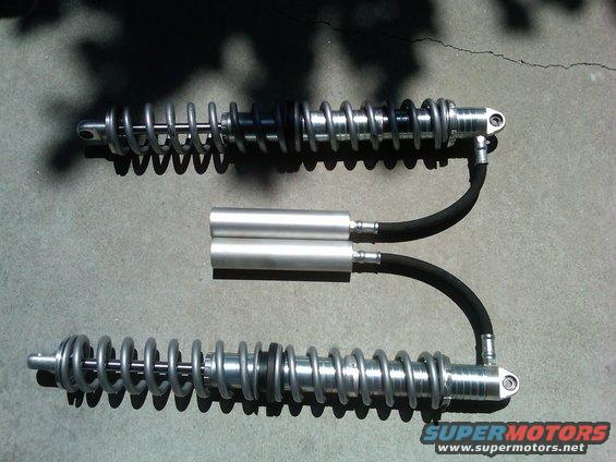 Two silver shock absorbers with black hoses and reservoirs sit on a concrete surface