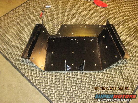 Black metal skid plate with many rivets, lying on a patterned mat. A screwdriver and tool are nearby