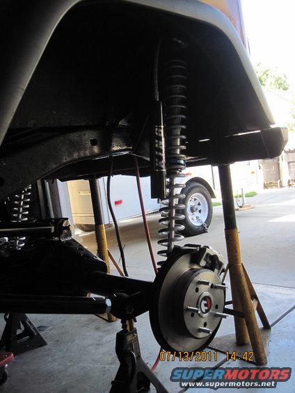 Close-up of a Jeep's front suspension and brake assembly, supported by jack stands. A trailer is visible in the background
