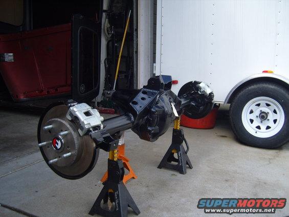 Jeep rear axle assembly with disc brakes on jack stands, red Jeep body in background, white trailer wheel to the right