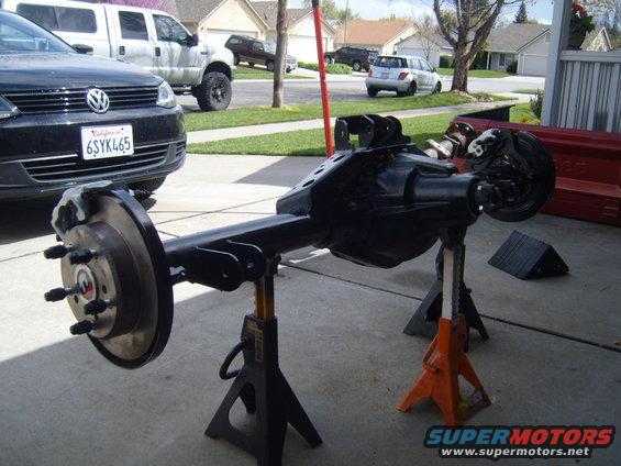 Rear axle assembly on jack stands in driveway, with Volkswagen and pickup truck in background