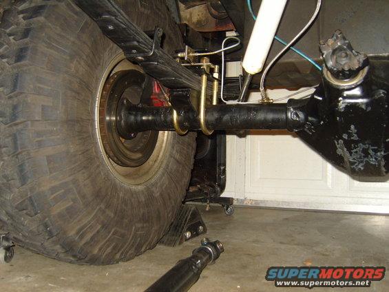 Close-up of a Jeep's rear axle, leaf springs, and large tire. A shock absorber is visible, and a driveshaft lies on the ground
