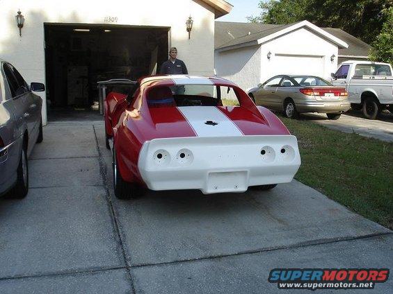 1976 Chevrolet Corvette with red body and white racing stripes, rear view. Another car and truck are parked nearby