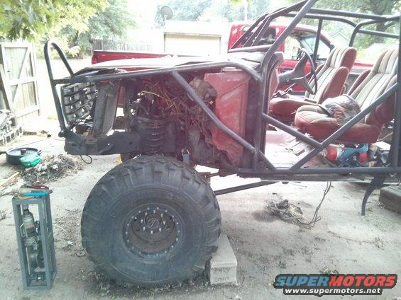 Custom off-road buggy frame with large tires, exposed engine, and bucket seats. Another red truck is visible in the background